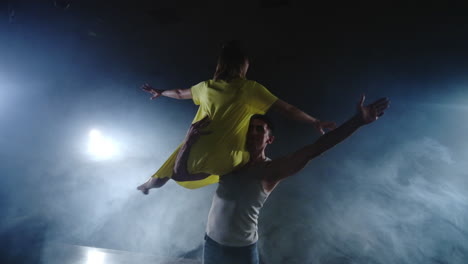 Zoom-camera-two-modern-ballet-dancers-are-dancing-on-stage-in-smoke-a-man-raises-his-partner-in-his-arms-and-rotates-in-the-air.-The-musical-uses-circus-acrobats-dancers.